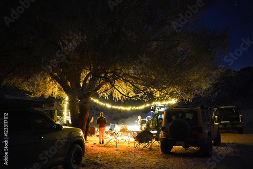 A cozy desert campsite at night, illuminated by string lights wrapped around a tree. Off-road vehicles and camping chairs surround a warm fire, creating a peaceful and adventurous outdoor atmosphere.