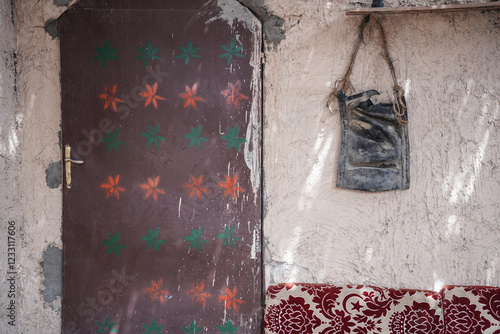 Traditional painted door with colorful star motifs beside a textured beige wall and an old leather bag hanging above patterned seating. A rustic cultural scene reflecting heritage and handcrafted desi