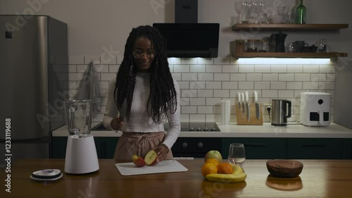 African black woman in glasses and casual outfit that cooking healthy smoothie with blender and cutting fruits in a modern kitchen at home. photo