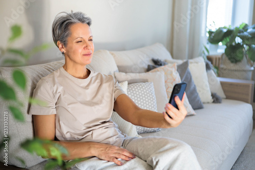 Mature beautiful middle-aged woman with gray hair and poor eyesight sits on the couch