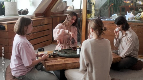 A girl is conducting a tea ceremony for a group of people. She is pouring freshly brewed tea into ceramic cups. The girls are drinking tea and talking.