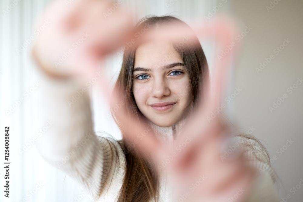 Fototapeta premium Girl framing her face with her hands in a heart shape. A young girl with brown hair smiles gently while framing her face with her hands in a heart shape, standing against a softly lit neutral