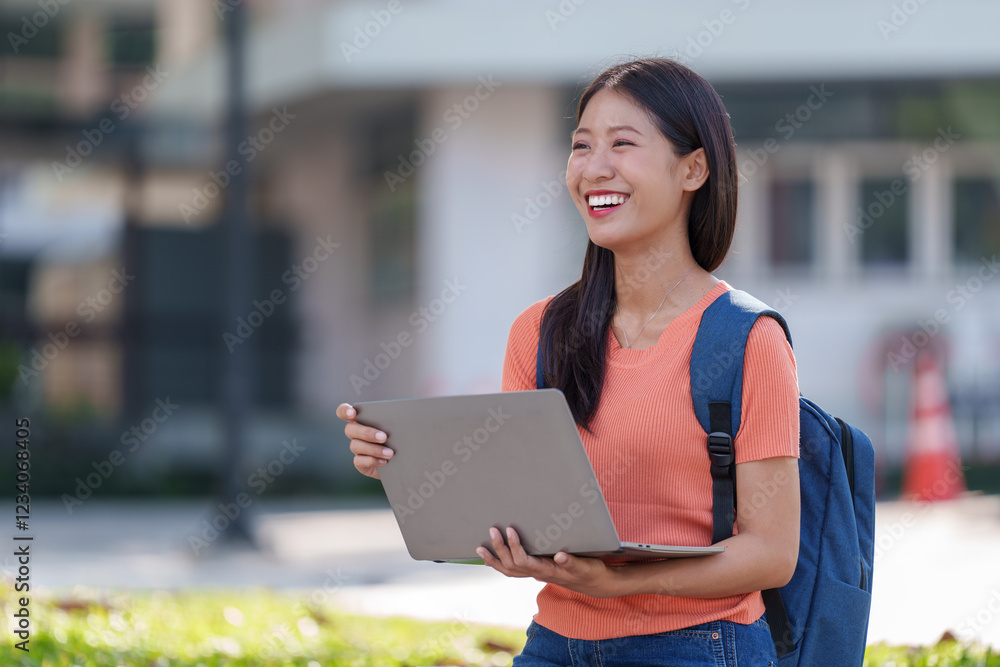 Fototapeta premium Cheerful university student is holding a laptop and smiling while walking on campus, enjoying the pleasant weather and academic atmosphere