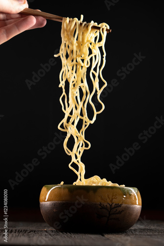 Steaming hot noodles being lifted from a  bowl by a pair of wooden chopsticks. The golden-yellow noodles cascade down, releasing wisps of steam that curl. Instant noodles on dark background. Food conc