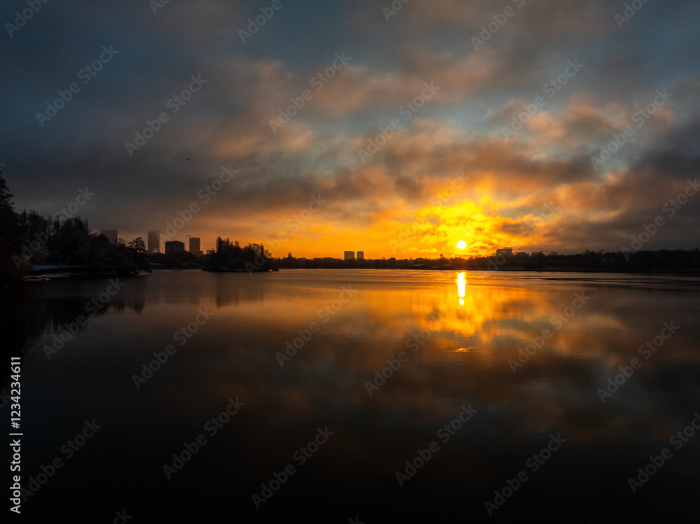 A serene winter sunrise over Bucharest, showcasing a glowing sun reflecting on the calm water surface, framed by misty clouds and silhouetted cityscape. Bucharest aerial winter photo over Herastrau