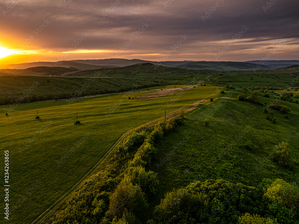 Dramatic sunset over a Transylvanian village surrounded by rolling green hills, capturing the essence of rural life and the charm of this historic region. Aerial photo form Transylvania, Romania.