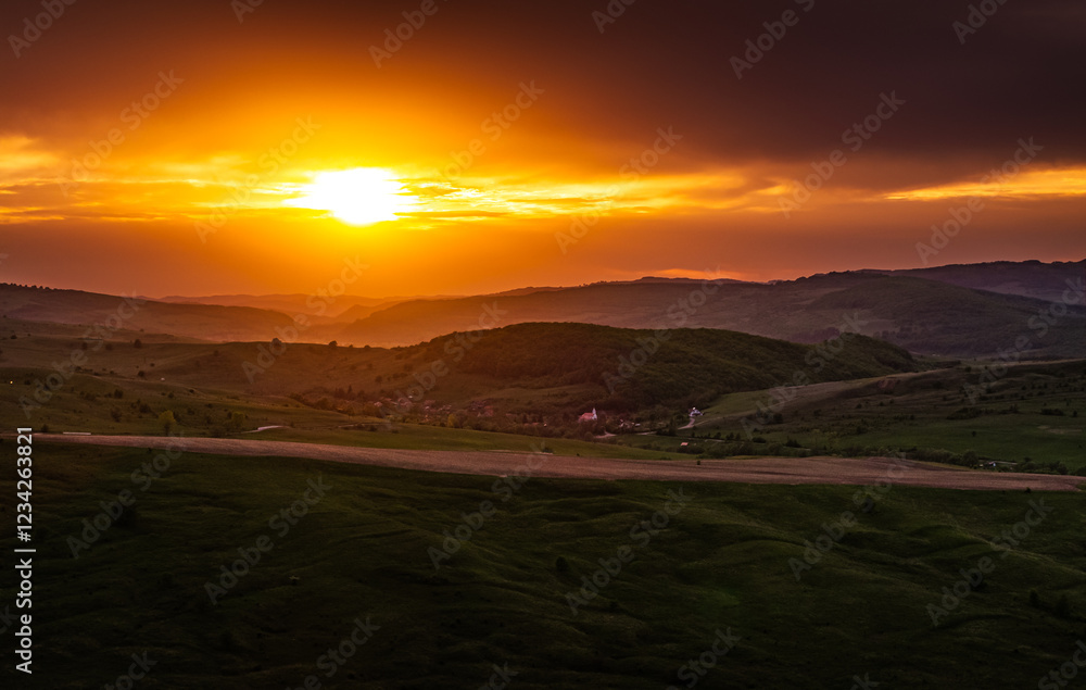 Dramatic sunset over a Transylvanian village surrounded by rolling green hills, capturing the essence of rural life and the charm of this historic region. Aerial photo form Transylvania, Romania.