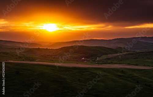 Obraz na płótnie Dramatic sunset over a Transylvanian village surrounded by rolling green hills, capturing the essence of rural life and the charm of this historic region. Aerial photo form Transylvania, Romania. w salonie
