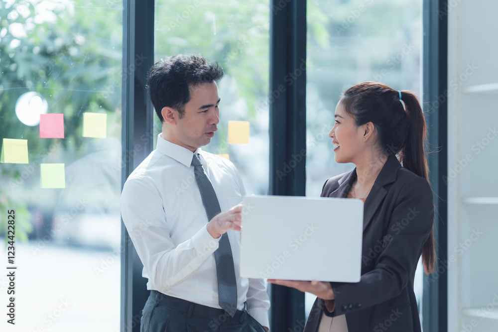 Business Collaboration: A man and woman in business attire stand next to a window and discuss ideas while holding a laptop, creating a professional, focused atmosphere for teamwork. Business Collaboration: A man and woman in business attire stand next to a window and discuss ideas while holding a laptop, creating a professional, focused atmosphere for teamwork.