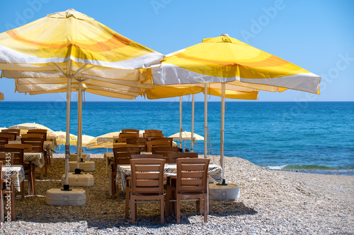 Fototapeta Naklejka Na Ścianę i Meble -  Relaxing beachside cafe scene. Wooden tables and chairs under vibrant yellow umbrellas, overlooking a stunning turquoise sea. Perfect for summer travel, vacation, and restaurant themes
