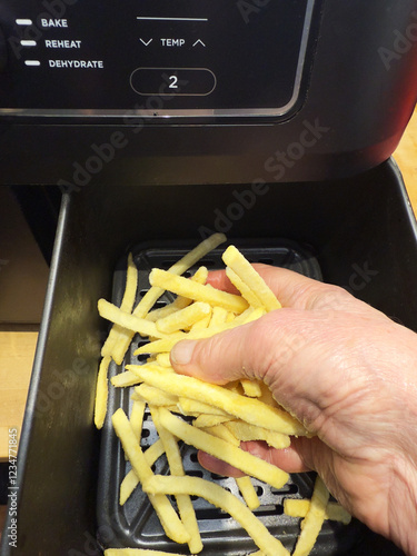 Closeup of a man’s hand placing frozen uncooked fries into the basket, bucket or drawer of an air fryer, ready to cook. 