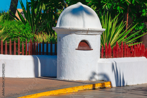 Small white historical guardhouse with a domed roof, set against lush green plants and a red wooden fence. Bright sunlight casts shadows on the pavement, enhancing Mediterranean architectural charm photo