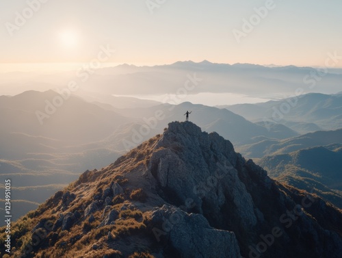 A silhouette of a person standing triumphantly on a rugged mountaintop at sunrise, with expansive views of misty valleys and distant peaks. photo