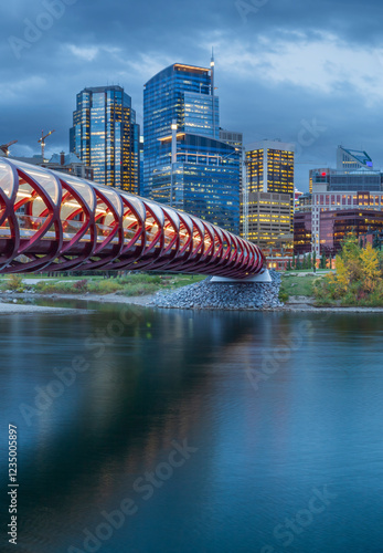 A panoramic view of the Peace Bridge in Calgary, Canada, at dusk. The red pedestrian bridge spans the Bow River, with the city's modern skyline and autumn foliage in the background.