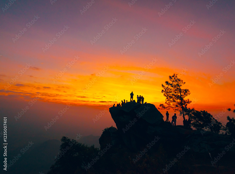 Obraz premium Silhouette of a group of people climbing on a large cliff. Colorful sky of orange, yellow, pink, and blue in the morning at sunrise. There are mountains and trees around with copy space on top.