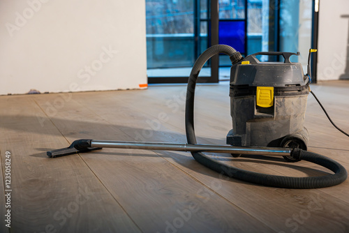 A heavy duty vacuum cleaner with yellow accents sits on a wooden floor indoors. Large glass doors reveal an outdoor area with a railing and blurred structures. photo