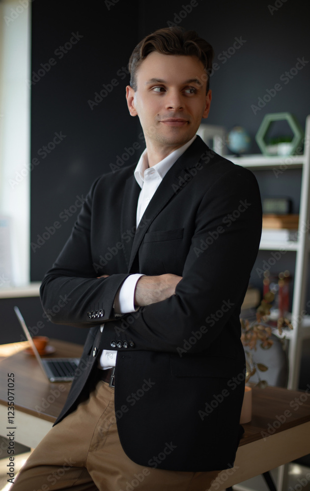 A confident businessman standing with arms crossed, dressed in a black suit and white shirt. The photo is taken in a modern office setting with a laptop and plants in the background. A confident businessman standing with arms crossed, dressed in a black suit and white shirt. The photo is taken in a modern office setting with a laptop and plants in the background.