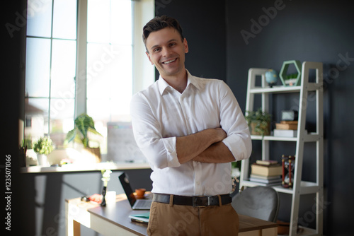 A confident man in a white shirt stands with arms crossed in a modern office space. He is smiling, looking off to the side, with plants, a laptop, and bookshelves in the background. photo