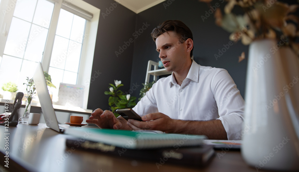 Fototapeta premium A young professional man wearing white shirt is sitting at a desk in a modern office. He is using a smartphone and a laptop, with plants and cup in the background, showing a focused working atmosphere