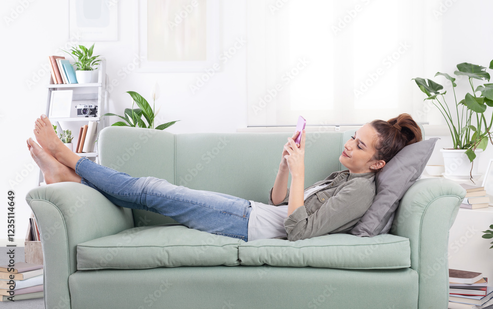 Young relaxed smiling woman lying on a cozy couch in a casual outfit, holding a smartphone and browsing messages, social media, and digital content Young relaxed smiling woman lying on a cozy couch in a casual outfit, holding a smartphone and browsing messages, social media, and digital content