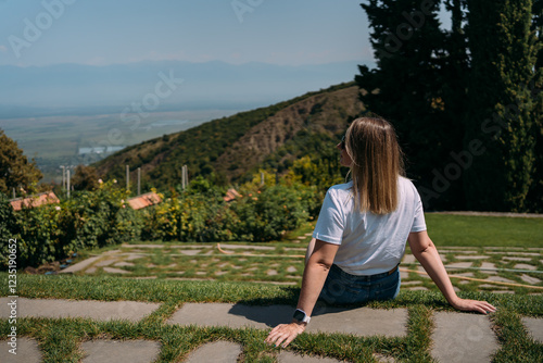 A smiling woman in a white T-shirt sitting on a grass outdoors with a scenic mountain view. view from the back photo