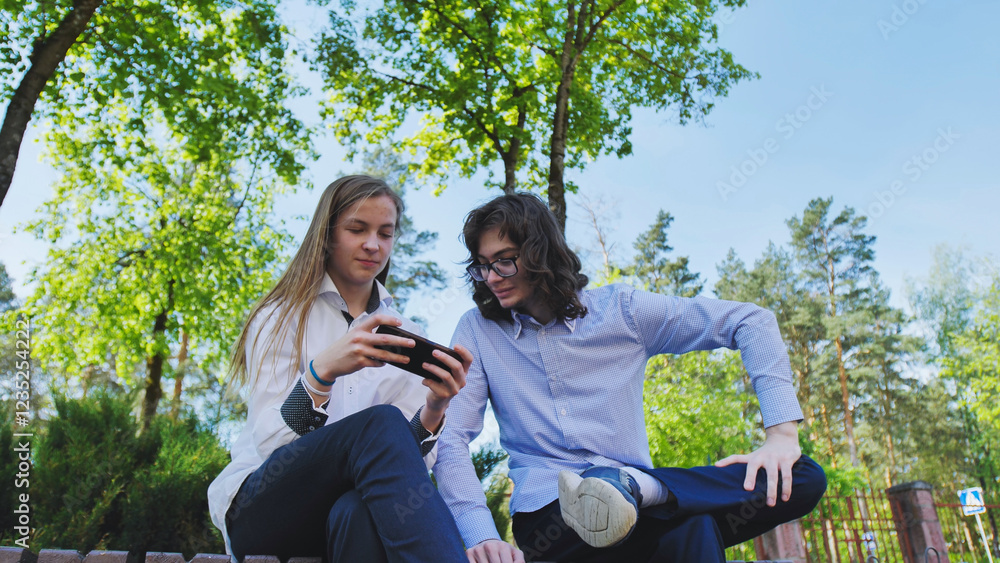 Two high school students are sitting on a bench in a park, looking at a smartphone and enjoying their school day Two high school students are sitting on a bench in a park, looking at a smartphone and enjoying their school day