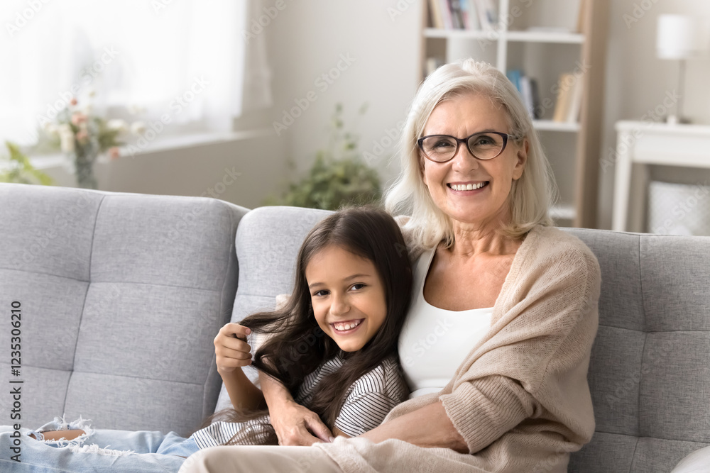 Warm relationship. Smiling middle aged grandmother hold in arms little girl tween age granddaughter look at camera. Granny and grandchild relax at home posing for family portrait on cozy sofa together Warm relationship. Smiling middle aged grandmother hold in arms little girl tween age granddaughter look at camera. Granny and grandchild relax at home posing for family portrait on cozy sofa together