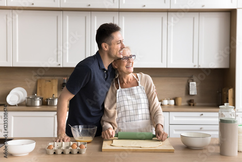 Dreamy young son tender hug shoulders of smiling retired mum baking buns at domestic kitchen look away together with beloved parent. Happy family aged mom grown up kid remember good day imagine future photo