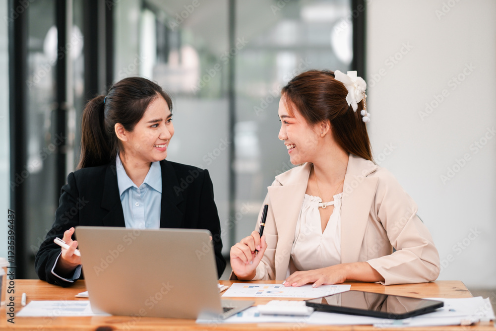 Two businesswomen discussing work at a desk with a laptop, showcasing teamwork and collaboration in a modern office environment. Two businesswomen discussing work at a desk with a laptop, showcasing teamwork and collaboration in a modern office environment.