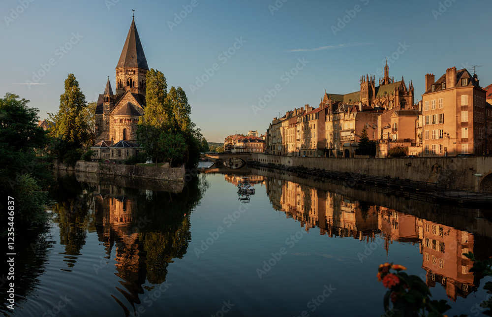 Obraz premium Historic riverside cityscape with a stone church, tree-lined banks, and reflective calm water illuminated by golden evening light.