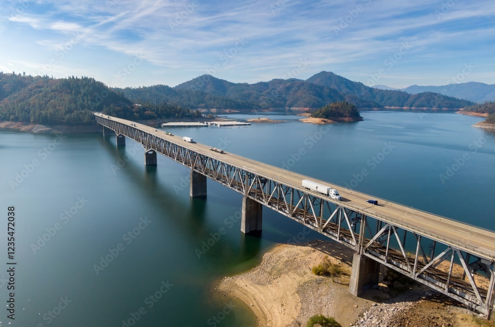 Elevated view of a steel bridge spanning Shasta Lake, with vehicles traversing the roadway. A scenic landscape of mountains and water surrounds the structure. Lakehead, California, USA Elevated view of a steel bridge spanning Shasta Lake, with vehicles traversing the roadway. A scenic landscape of mountains and water surrounds the structure. Lakehead, California, USA