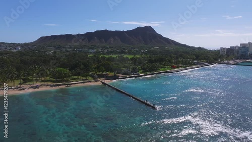 A slow aerial pan of Waikiki Beach with Diamond Head in the background, turquoise waters, a seawall, and modern buildings along the lush shoreline. photo