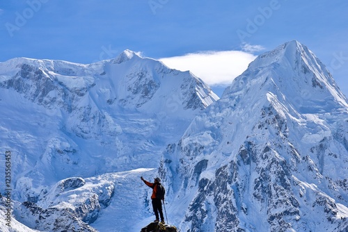 Fotografie A trekker in a red jacket stands triumphantly on a rocky peak near Kanchenjunga Base Camp South, Nepal, surrounded by the majestic snow-covered Himalayas under a clear blue sky