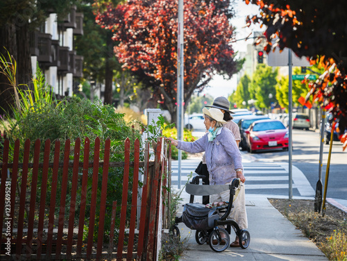 Senior woman walking using a mobility walker on the pedestrian walkway, checking the plants  outside the retirement apartment. California.