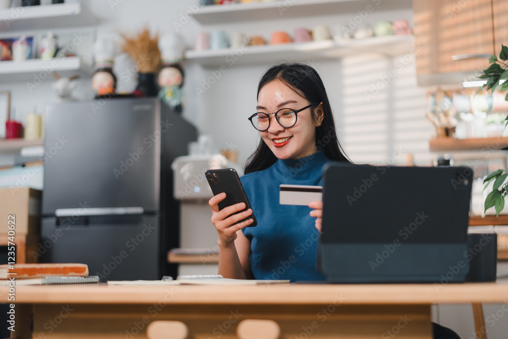 woman smiling while using smartphone and holding credit card, sitting at desk with tablet in cozy kitchen woman smiling while using smartphone and holding credit card, sitting at desk with tablet in cozy kitchen