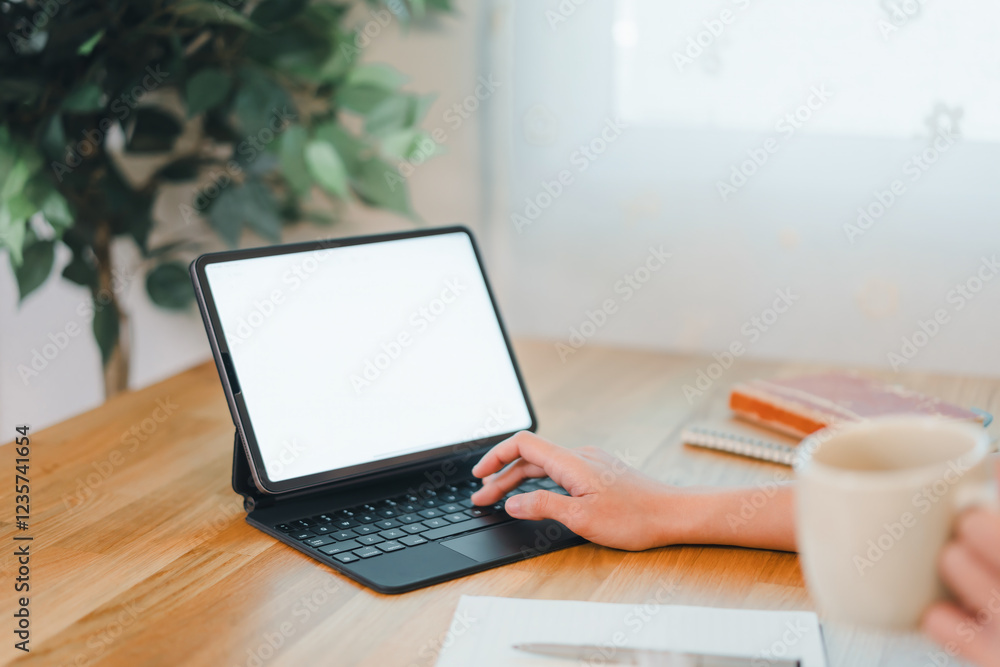 Fototapeta premium person working on tablet with keyboard case on wooden desk, holding coffee cup, surrounded by notebooks and plant