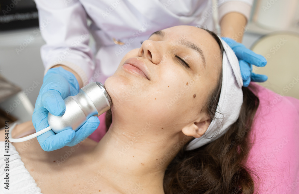 Naklejka premium Woman is getting a facial treatment with a RF lifting device on her face. The woman is wearing a white bandana and a blue glove
