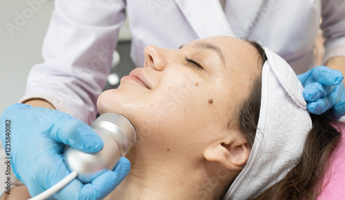 Woman is getting a facial treatment with a RF lifting device on her face. The woman is wearing a white bandana and a blue glove photo