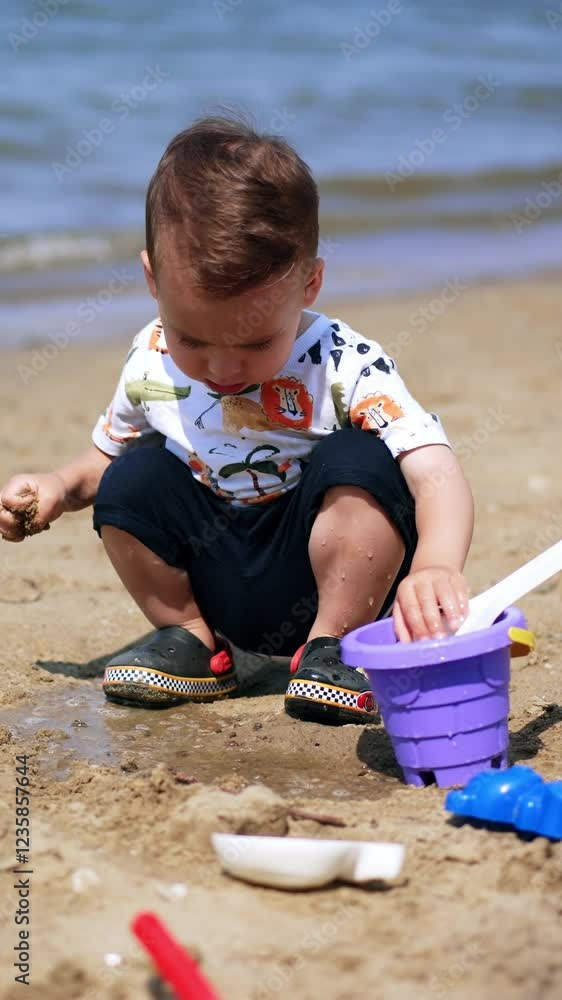 Adorable toddler boy plays with toys at the seashore. Kid pours some water from his bucket on the sand. Vertical video Adorable toddler boy plays with toys at the seashore. Kid pours some water from his bucket on the sand. Vertical video