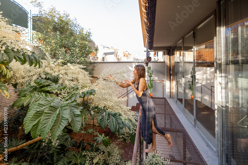 A woman in a minimalist dress leans on a railing, enjoying lush greenery on a modern balcony. The soft sunlight and natural ambiance create a peaceful retreat, blending urban living with nature