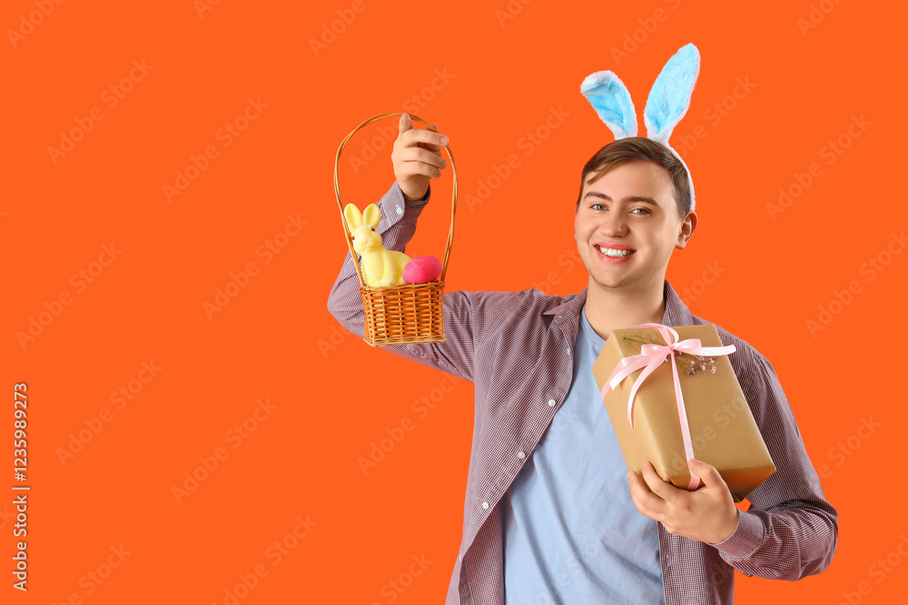 Happy young man in bunny ears with gift box and wicker basket on orange background. Easter celebration Happy young man in bunny ears with gift box and wicker basket on orange background. Easter celebration