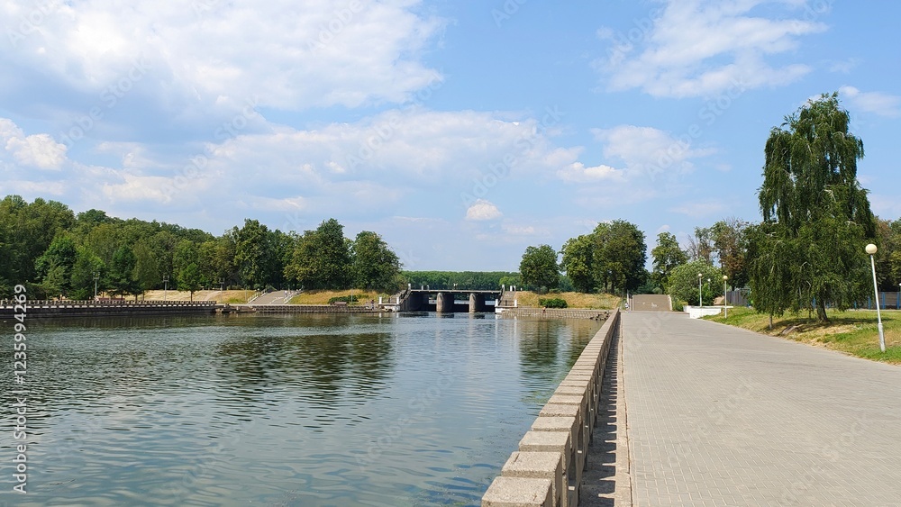 The banks of the river flowing through the city are concreted, with tile walkways and light poles along them. A dam blocks the river. Trees grow on the grassy lawns. Sunny summer weather and blue sky The banks of the river flowing through the city are concreted, with tile walkways and light poles along them. A dam blocks the river. Trees grow on the grassy lawns. Sunny summer weather and blue sky
