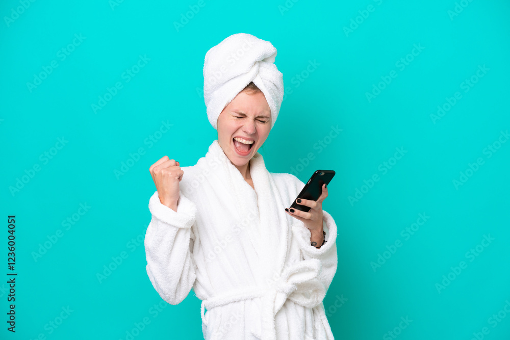 Young woman in a bathrobe isolated on blue background with phone in victory position Young woman in a bathrobe isolated on blue background with phone in victory position
