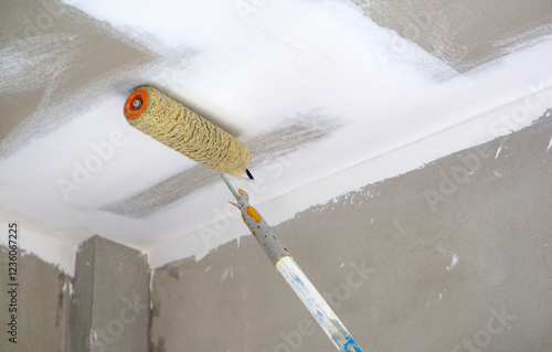 A paint roller is being used to apply a fresh coat of white paint on the ceiling of a newly renovated room. The wall have a smooth finish, indicating recent update.
