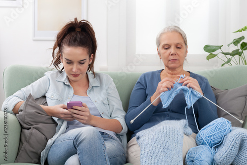 Fotografi At home on the couch, a Millennials daughter uses her smartphone to browsing while her senior mother Boomer sits nearby knitting
