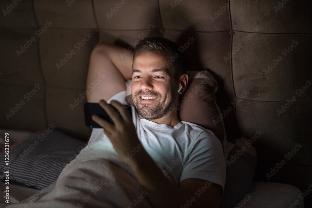Fototapeta premium Young man is watching shows on cellphone late at night in dark bedroom.