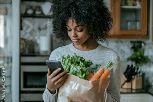 A woman in a kitchen looking at her cell phone while holding a bag of food