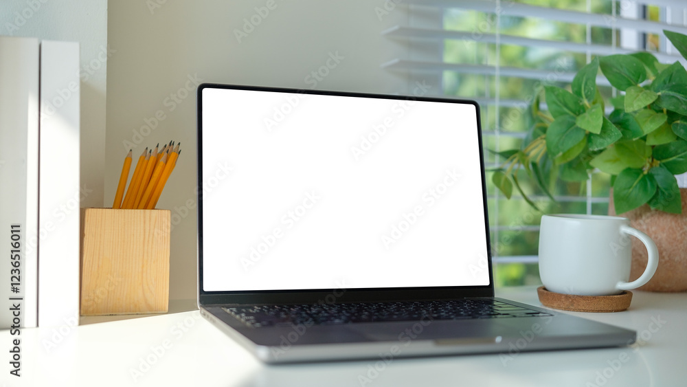 Fototapeta premium Laptop with blank screen, pencil holder and coffeecup on white desk, accented by natural light streaming through window blinds.