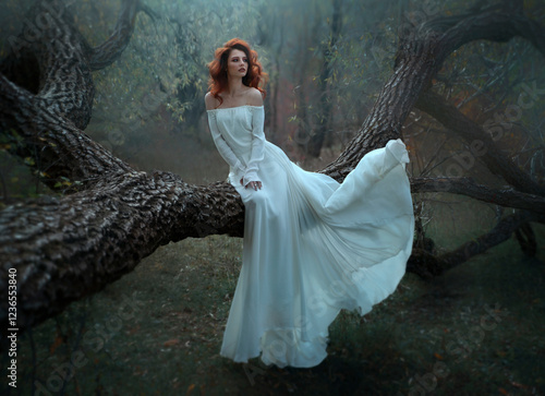 Beautiful red-haired woman in long white sitting on a tree in the forest.