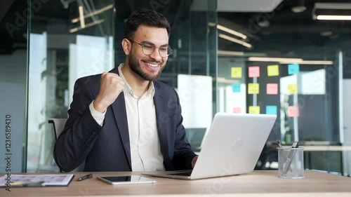Satisfied happy businessman in a formal suit received great news on laptop while sitting at workplace in business office. Smiling glad handsome entrepreneur reads positive good message on the computer